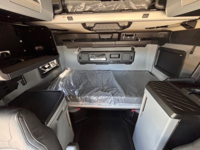Cab interior of a 2026 Mack PR64T76H showing sleeper area with forward control panel, storage compartments, and plastic-covered bed.
