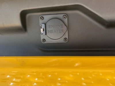 Engine heater access panel with two screws on a Mack PR64T76H truck, yellow surface below. The text ‘ENGINE HEATER’ is visible on the panel.
