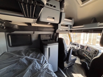 Interior of a 2026 Mack PR64T76H sleeper cab showing cockpit with steering wheel and dashboard, bunk area with overhead storage and cabinets, and protective cover on the bed.