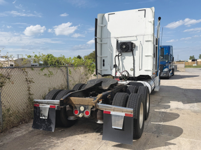 Truck rear view of a 2015 Freightliner Coronado with fifth-wheel and dual rear axles, blue cab in distance, white sleeper with satellite antenna on back, chain/link hitch area.