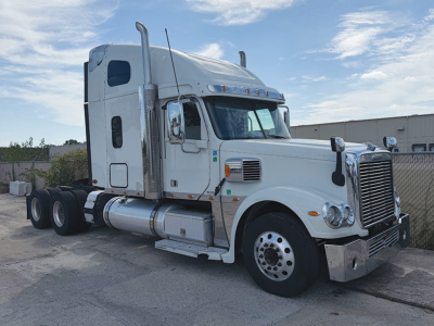 Truck cab and chassis showing a white 2015 Freightliner Coronado conventional sleeper tractor with chrome exhaust stacks, large grille, fuel tanks, and dual rear axles.