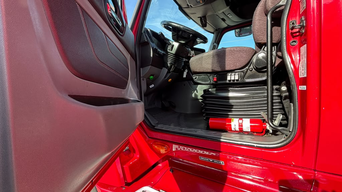 Truck cab interior of a 2022 International LT625 with red exterior, showing driver seat, dashboard, steering wheel, and a fire extinguisher mounted near the door.
