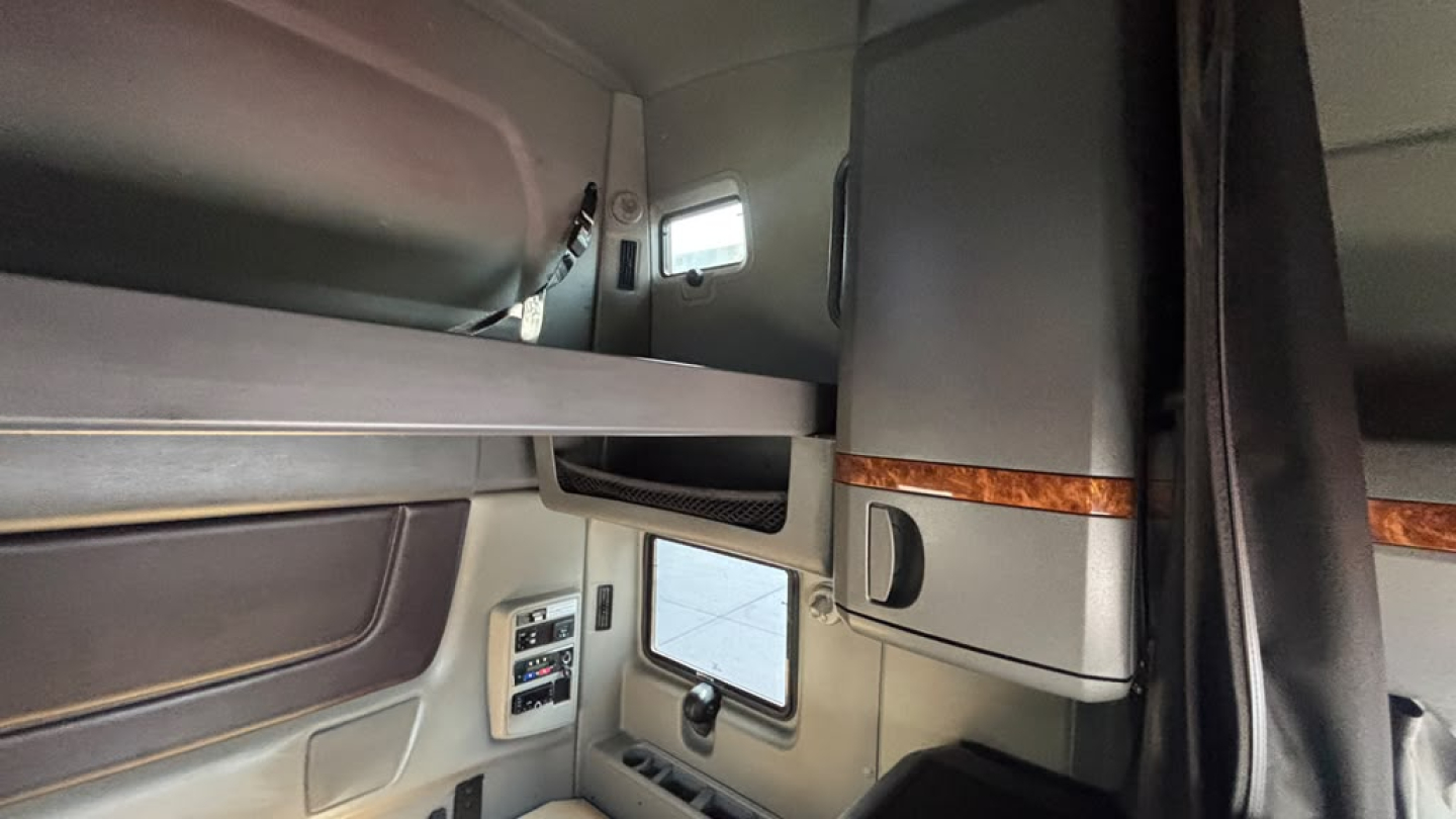 Cab interior of a 2022 International LT625 sleeper showing overhead bunk area, storage cabinet with wood trim, side window, and control panel.