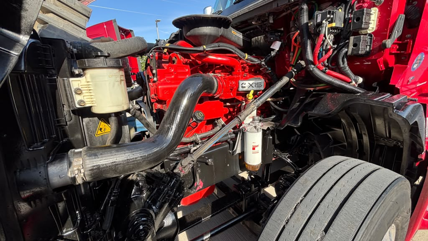 Truck engine bay showing red International LT625 with drivetrain components, hoses, and air filter housing visible.
