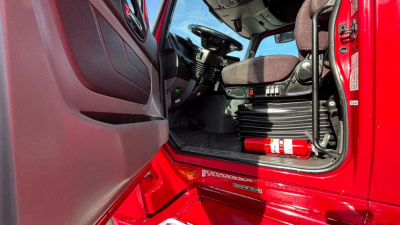 Truck cab interior showing red exterior, driver's seat, dashboard, and a red fire extinguisher mounted near the door.