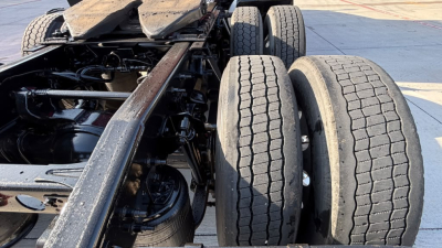 Three-axle heavy truck chassis with dual rear axles and dual tires; underside view showing frame rails, suspension components, and drivetrain.