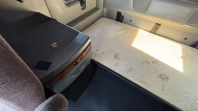 Truck interior showing a flat sleeping berth or storage area with a light-colored surface and a brown seat near a cabinet; a small black case sits on a panel.