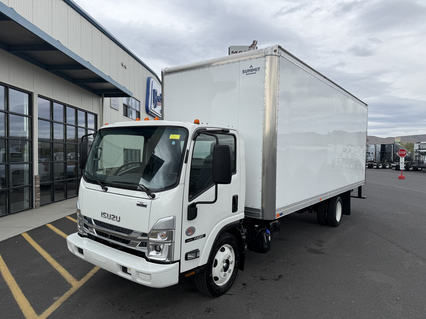 Isuzu NQR box truck with cab in front, white cab and large white cargo box mounted on dual rear wheels, dealership lot in background.