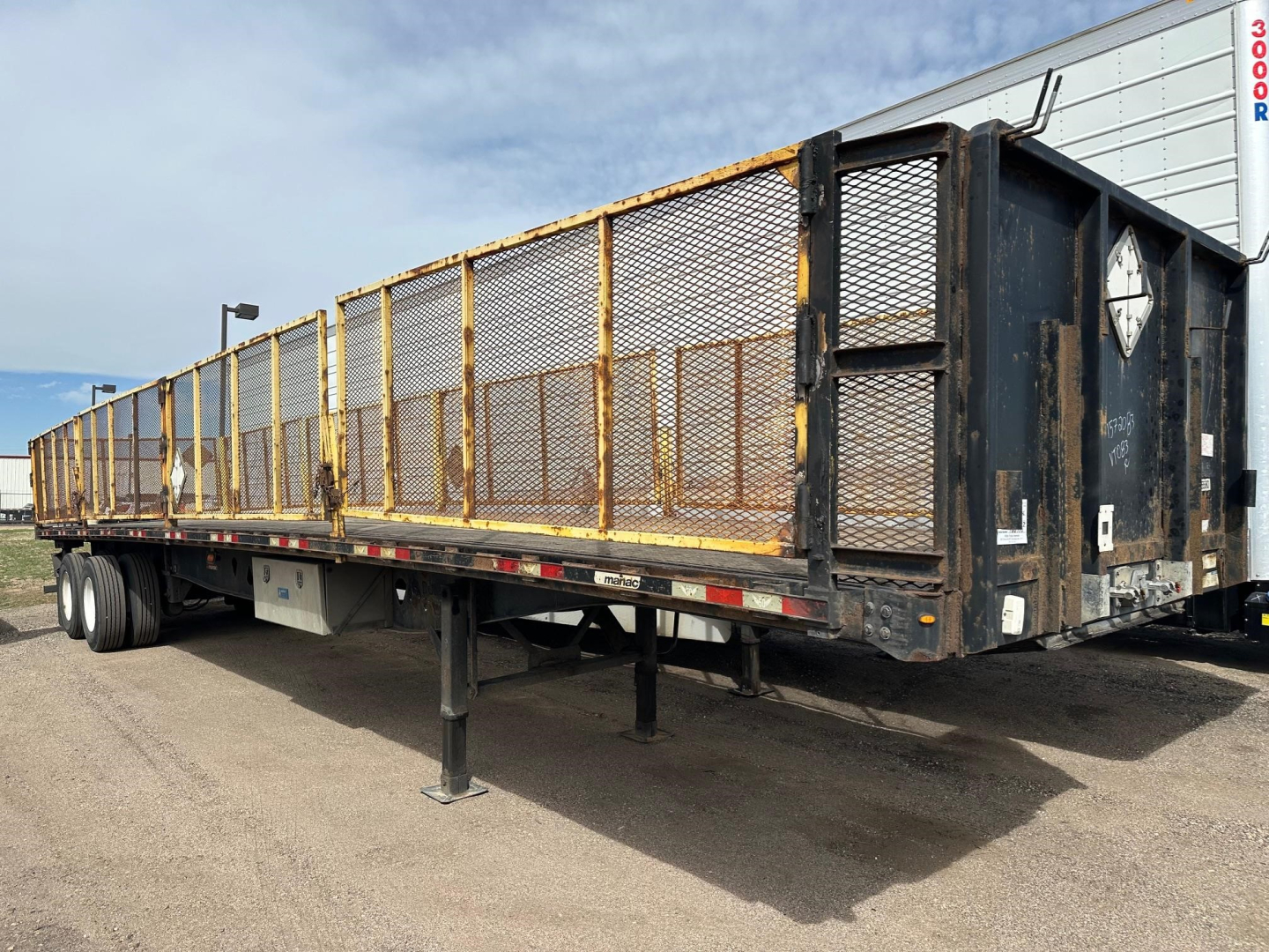 Manac flatbed trailer with closed tandem air ride, black frame and yellow mesh side gates, side view showing stake pockets and rear hitch.