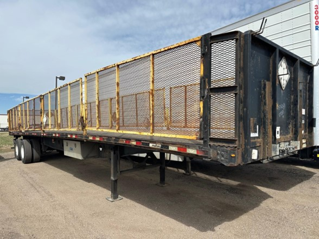 Manac flatbed trailer in service showing steel side rails and mesh panels, tandem axle configuration, air ride with drop legs extended.