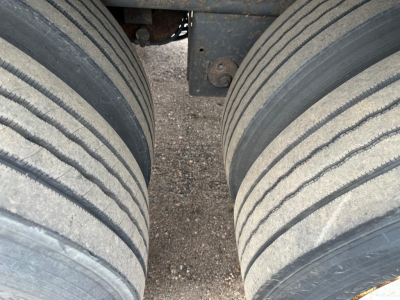 Trailer axle close-up showing dual rear tires on a Manac flatbed trailer, tandem air ride suspension visible, dirt/grit on ground