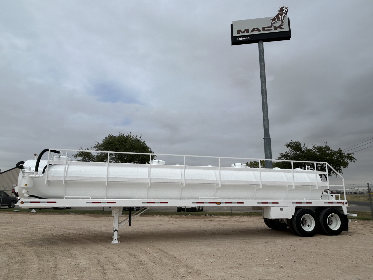Trailer in white with long cylindrical tank, dual rear axles, side rails, and support leg extended in gravel lot. Mack sign in background.