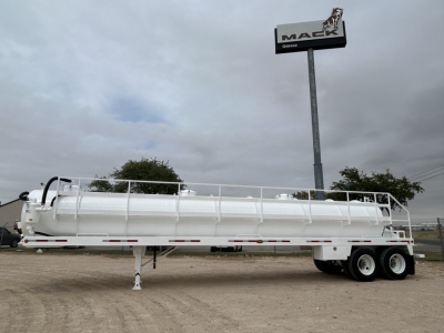 Trailer in white with long cylindrical tank, dual rear axles, side rails, and support leg extended in gravel lot. Mack sign in background.