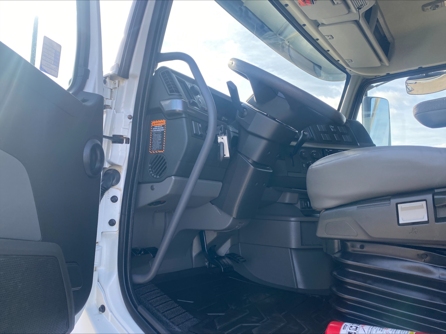Truck cab interior of a Volvo VHD; steering wheel, dashboard, and driver's seat visible; light entering from open door; dashboard controls and a vertical orange badge on the left side.