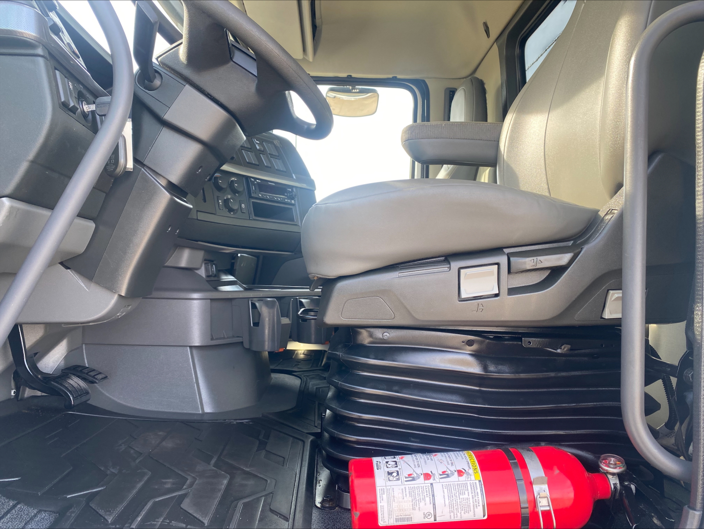 Interior of a Volvo VHD truck cabin showing steering wheel, dashboard, and driver's seat with a raised air-spring pedestal; a red fire extinguisher is visible on the floor near the pedals.