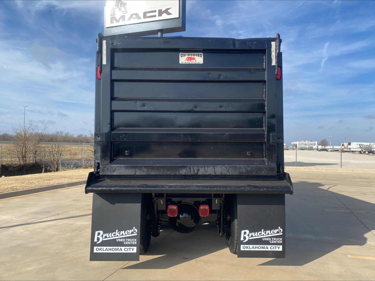 Back of a Volvo dump truck with a large black dump bed and red taillights; mud flaps show Buckner's Oklahoma City; clear blue sky.