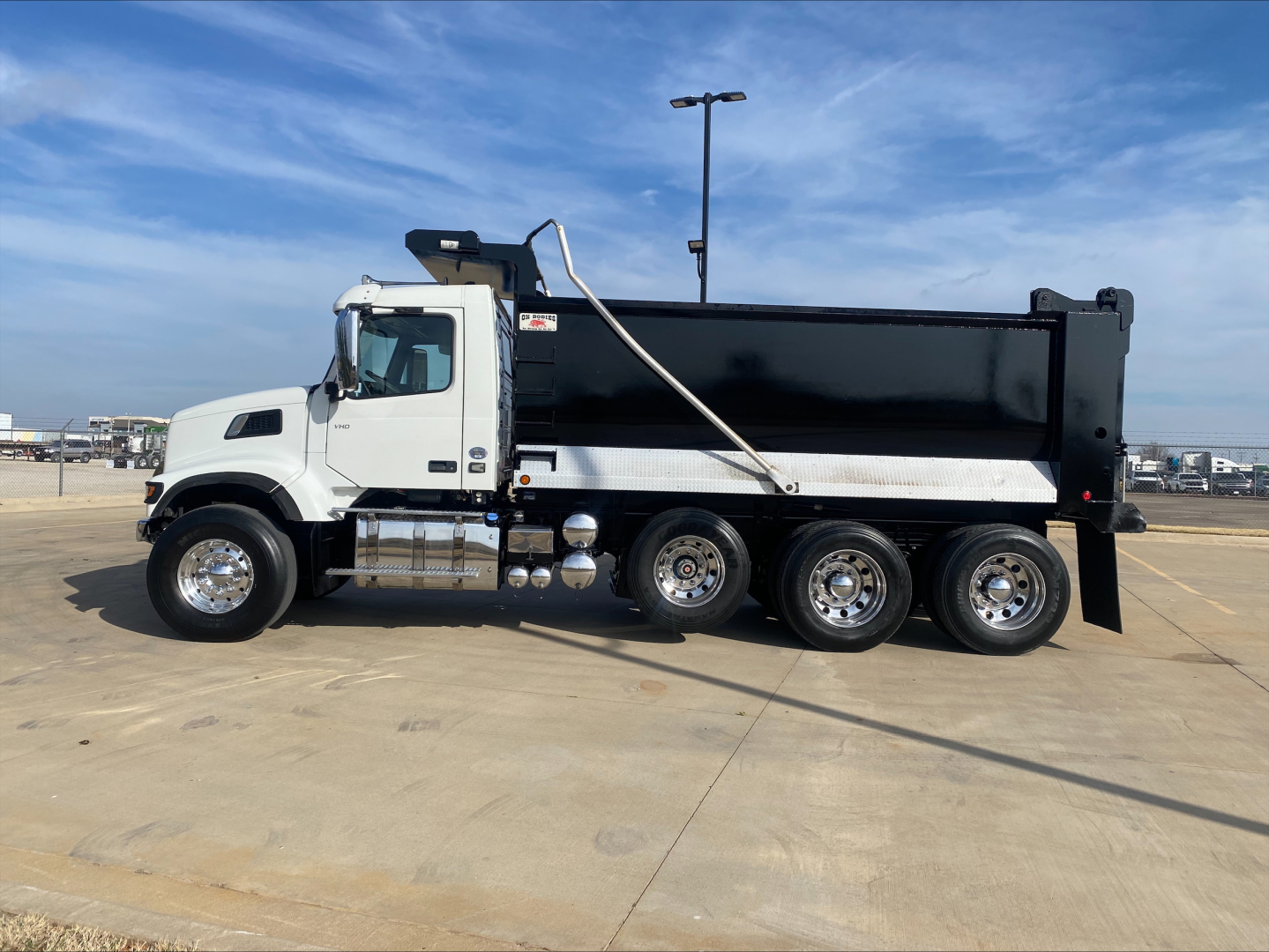 Dump truck in a parking lot with white cab and black dump bed; side view showing six wheels on the rear and chrome fuel tanks, with Volvo badge on door.