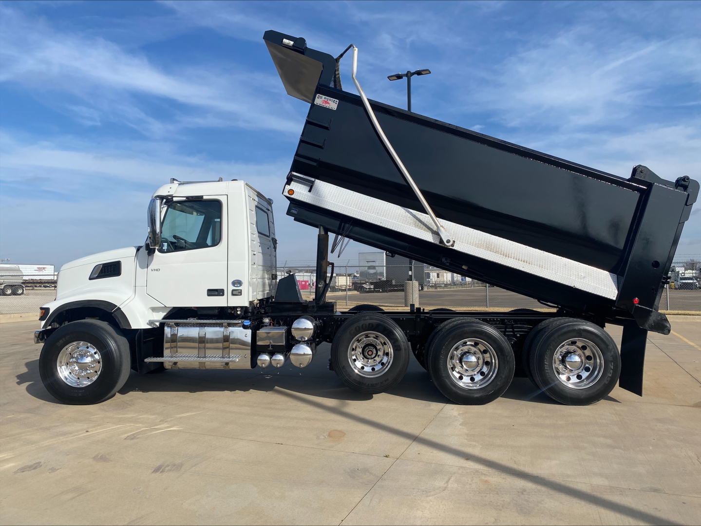 Truck side view showing a white Volvo dump truck with a raised black dump bed, multiple rear axles, chrome fuel tanks, and hydraulic lifting mechanism.