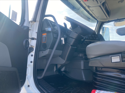Truck cab interior of a Volvo VHD; steering wheel, dashboard, and driver's seat visible; light entering from open door; dashboard controls and a vertical orange badge on the left side.