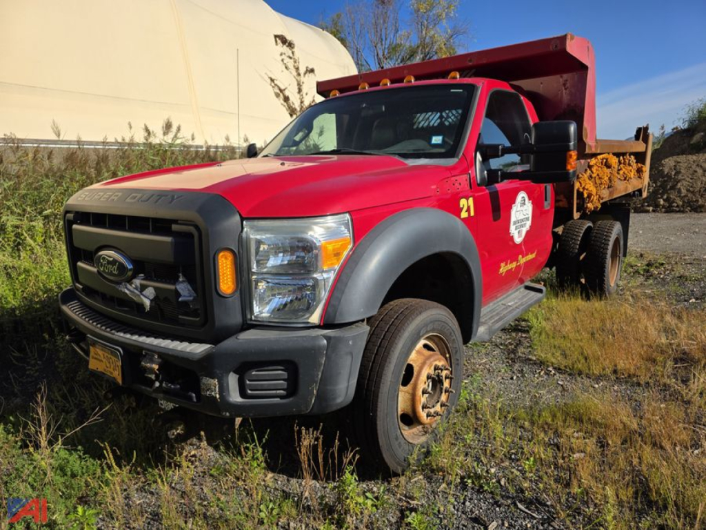 Ford F-550 Super Duty dump truck in red and black with a yellow dump body, front grille and bumper, VIN label on side window, parked on gravel with some vegetation