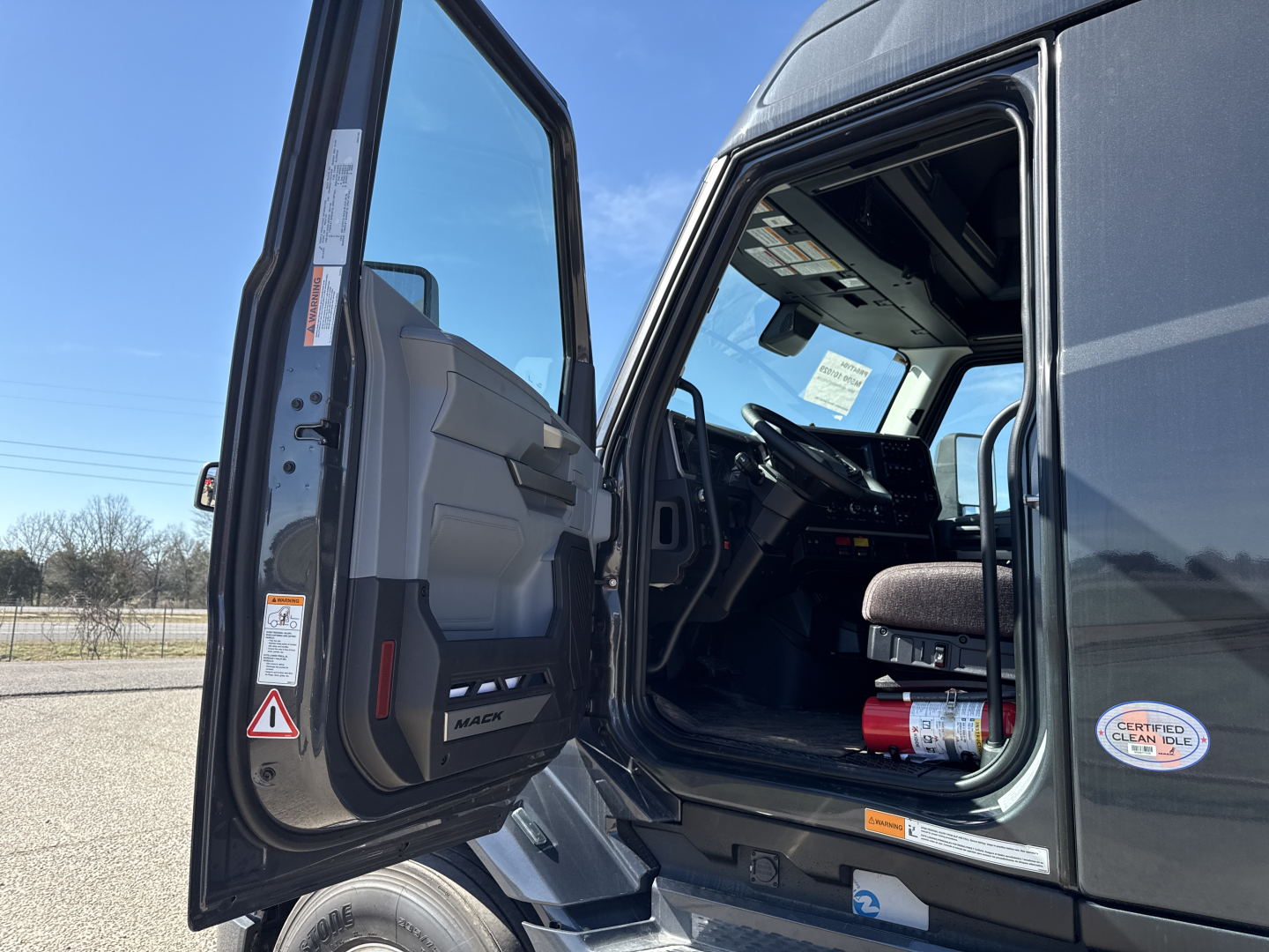 Truck cab interior of a 2026 Mack PR64T76H viewed from open driver door showing steering wheel, dashboard, and seat; VIN label and small sticker visible.