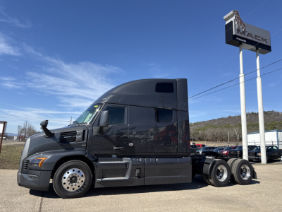 Truck side view of a new 2026 Mack PR64T76H conventional sleeper tractor in dark gray, parked on lot with Mack sign in background.