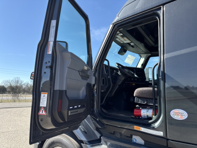 Truck cab interior of a 2026 Mack PR64T76H viewed from open driver door showing steering wheel, dashboard, and seat; VIN label and small sticker visible.