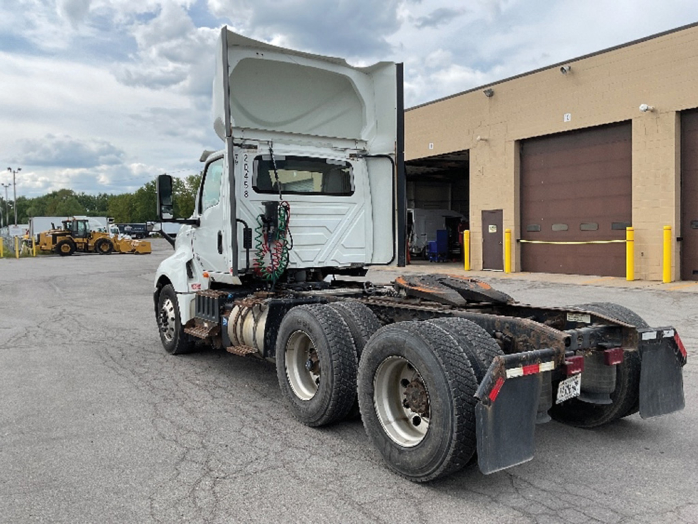 Truck chassis with cab lifted, rear dual-axle setup, various hoses and connections visible; white cab and flatbed area behind cab.