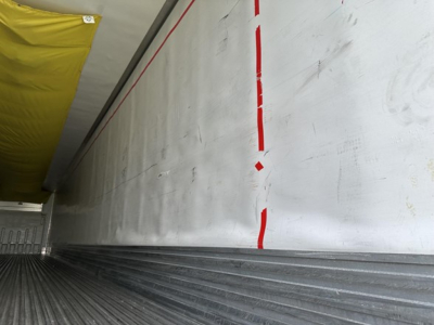 Trailer interior wall with a white metal surface, red vertical lines, and a yellow insulated cover along the top edge; ribbed floor and side wall details visible.