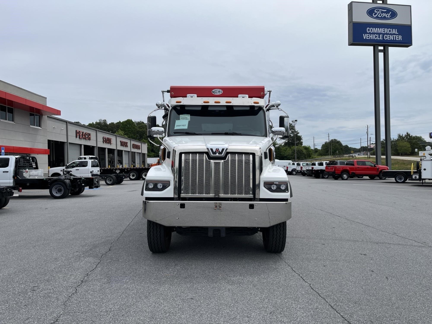 Front view of a 2027 Western Star 47X dump truck with large chrome grille, headlights, and orange light bar on cab, dealership lot in background.
