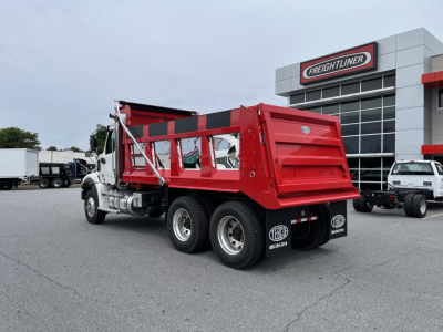 Dump truck rear angled view showing red dump bed, dual rear axles, and Freightliner dealership in background.