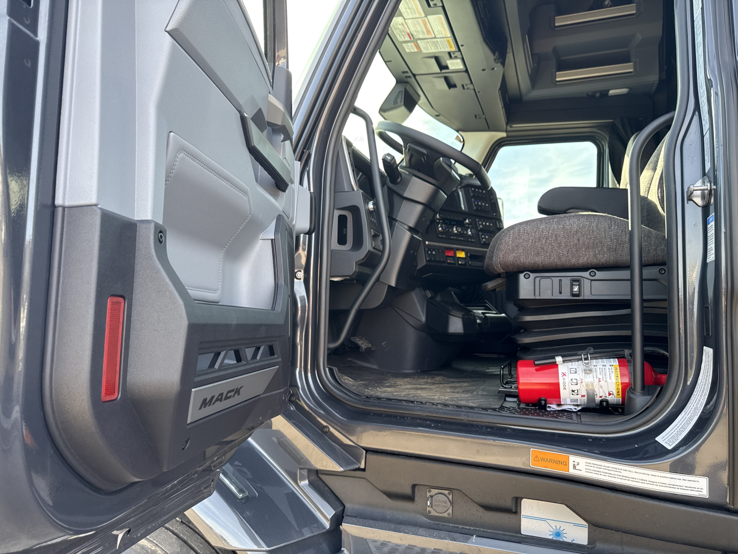 Truck cab interior of a 2026 Mack PR64T76H with open driver door, steering wheel, dashboard, and a red fire extinguisher in the cab floor.