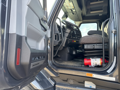 Truck cab interior of a 2026 Mack PR64T76H with open driver door, steering wheel, dashboard, and a red fire extinguisher in the cab floor.