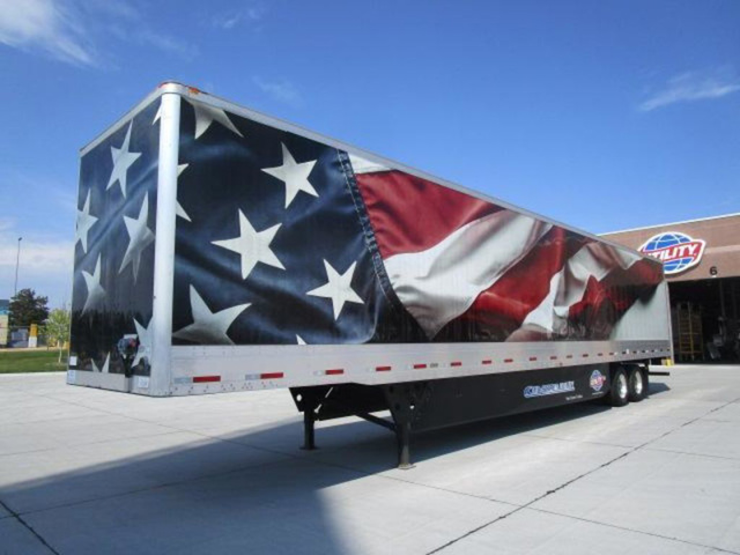 Trailer with American flag graphic, parked on lot with clear blue sky. Shows large box trailer with star-spangled artwork and dual-axle wheels.