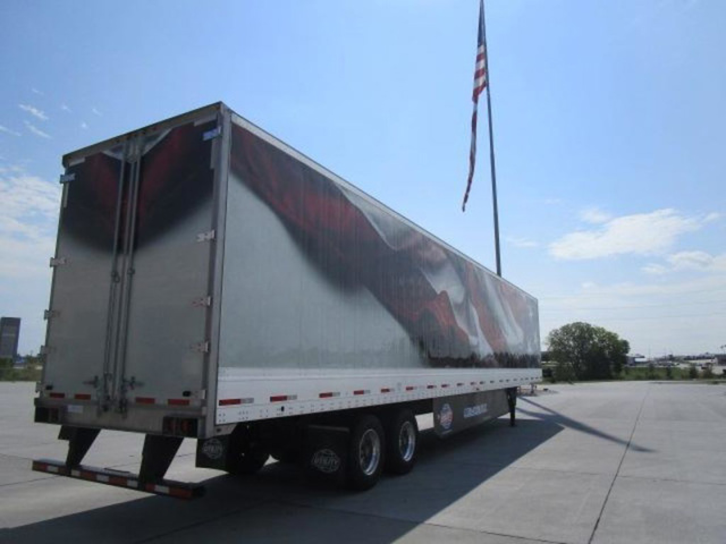 Trailer side view showing a large rectangular cargo trailer with triple axles and a reflective silver exterior; no legible label text visible on surface.