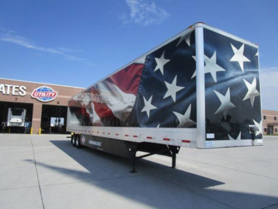 Utility van trailer with a large American flag design on the side, parked in a lot in front of a building with a sign. The trailer is a dry van with a pointed nose and dual rear axles visible under the wheel well.