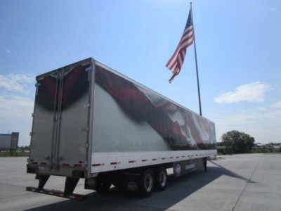 Trailer side view showing a large aluminum curtain-side trailer with three axles and an American flag in the background.