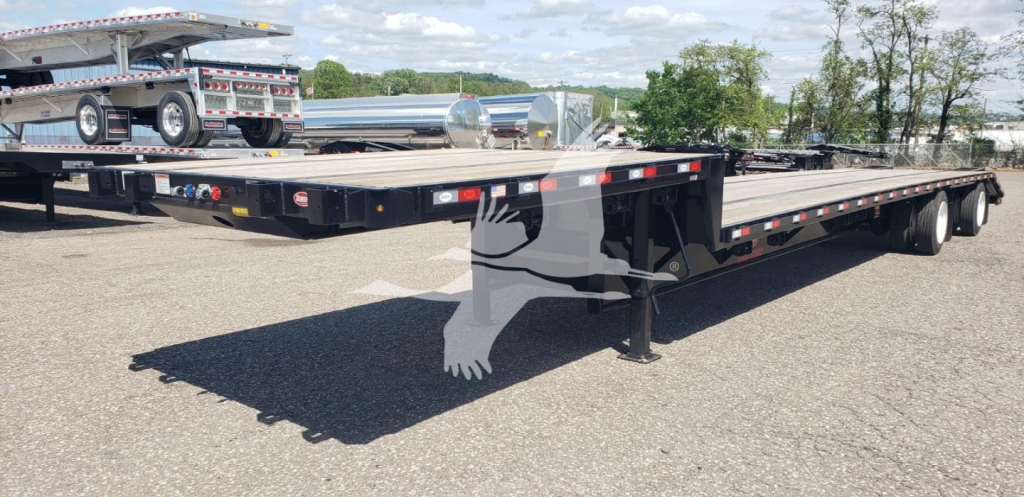 Flatbed trailer with drop-deck design, black frame, dual rear axles, and landing gear extended; storage and equipment visible in background.
