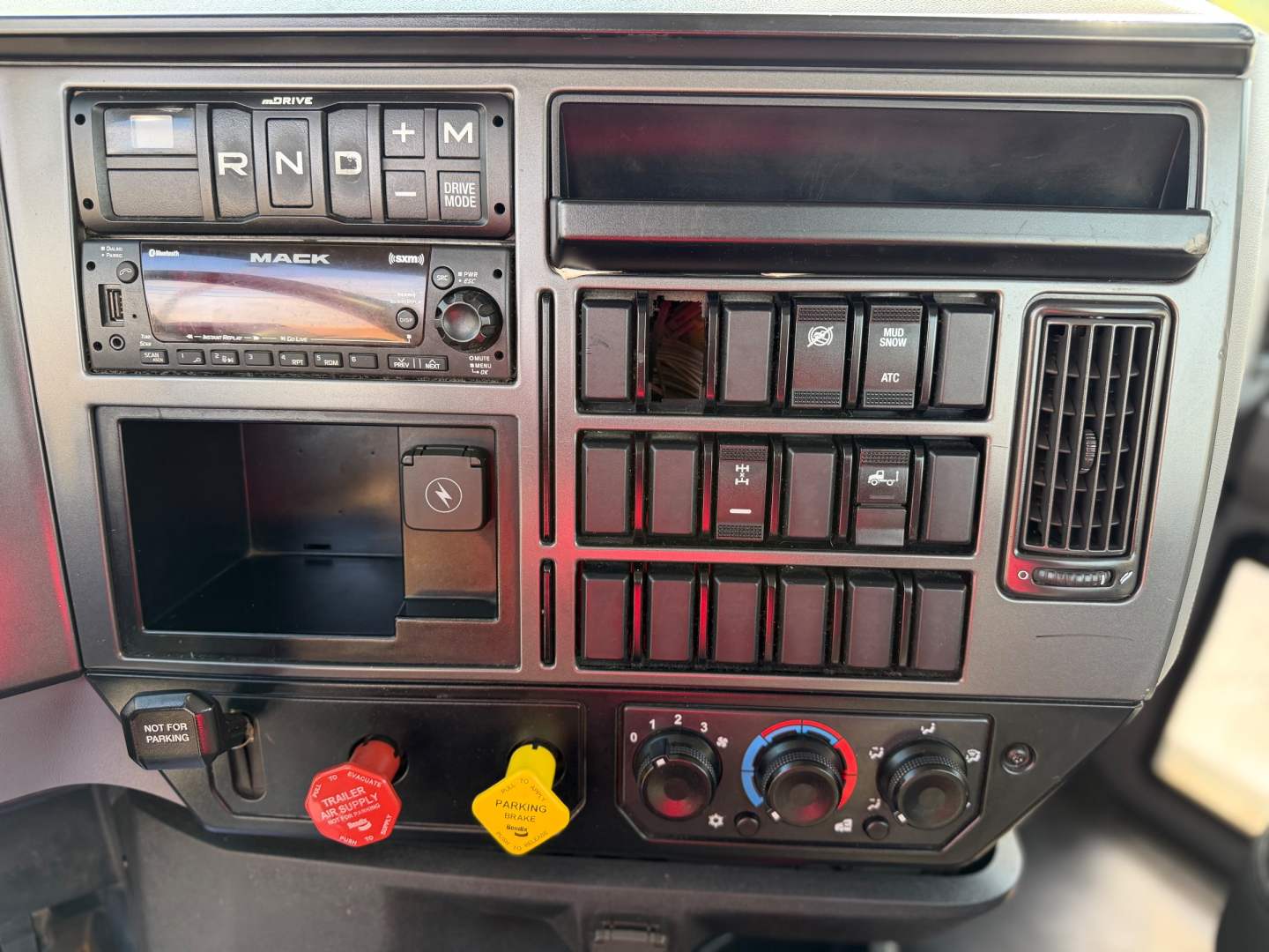 Dashboard of a Mack truck with controls, gauges, and switches. Visible text includes R N D P gear selector, Mack branded stereo, ignition area, red and yellow knobs, various switch blocks, and climate/auxiliary controls.