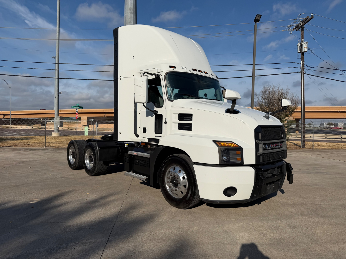 White Mack ANTHEM tractor with sleeper cab, front view, showing no visible text on side; truck sits on lot with power poles and fence in background.