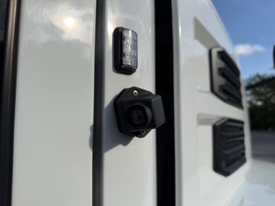 Camera-style rear-view of a white 2023 Mack Anthem 64T truck cab edge showing a small black camera mounted on the door frame near the bumper and a vertical marker light.