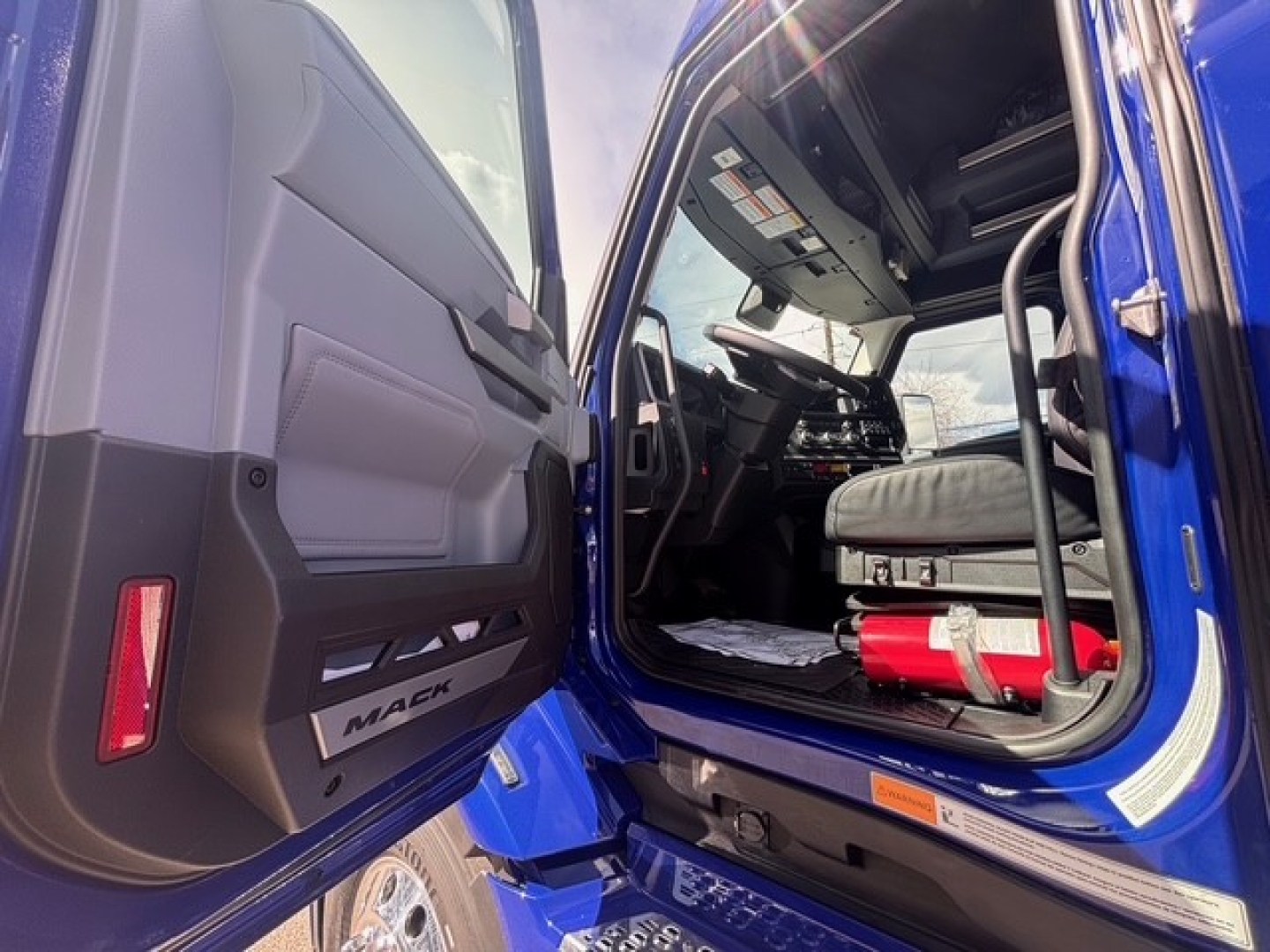 Truck cab interior of a new 2026 Mack PR64T76H sleeper with open door, steering wheel, dashboard, and blue exterior.