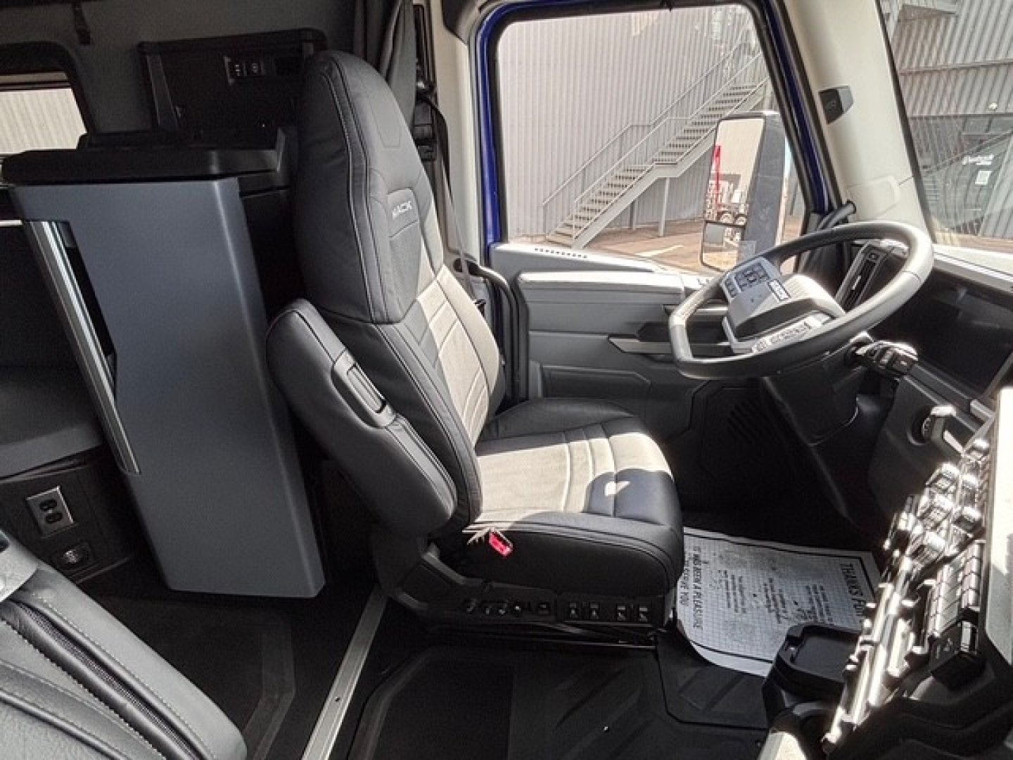 Truck cab interior of a new 2026 Mack PR64T76H showing driver seat, steering wheel, and dashboard area with a nearby fridge.