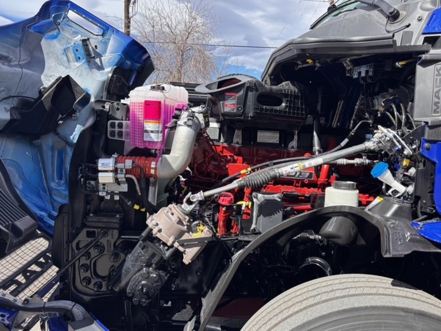 Mack PR64T76H engine bay view showing exposed red engine, pink coolant reservoir, and various hoses and wiring.