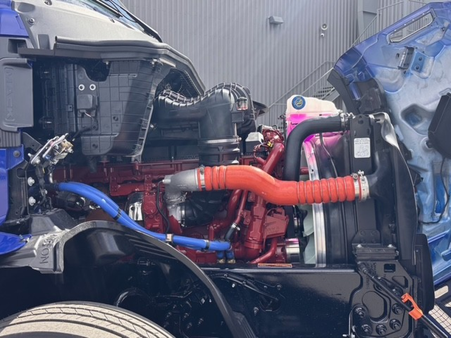 Mack PR64T76H engine bay with red engine components, blue hoses, and orange coolant hose visible between cab and radiator, mid-engine area.