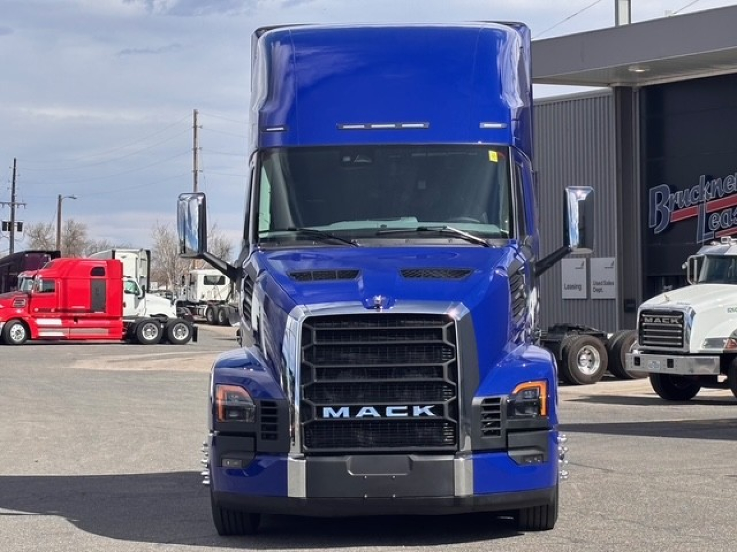 Truck front view showing Mack grille with large chrome bumper and blue cab; text 'MACK' on grille, truck hood, side mirrors, and blue sleeper cab.