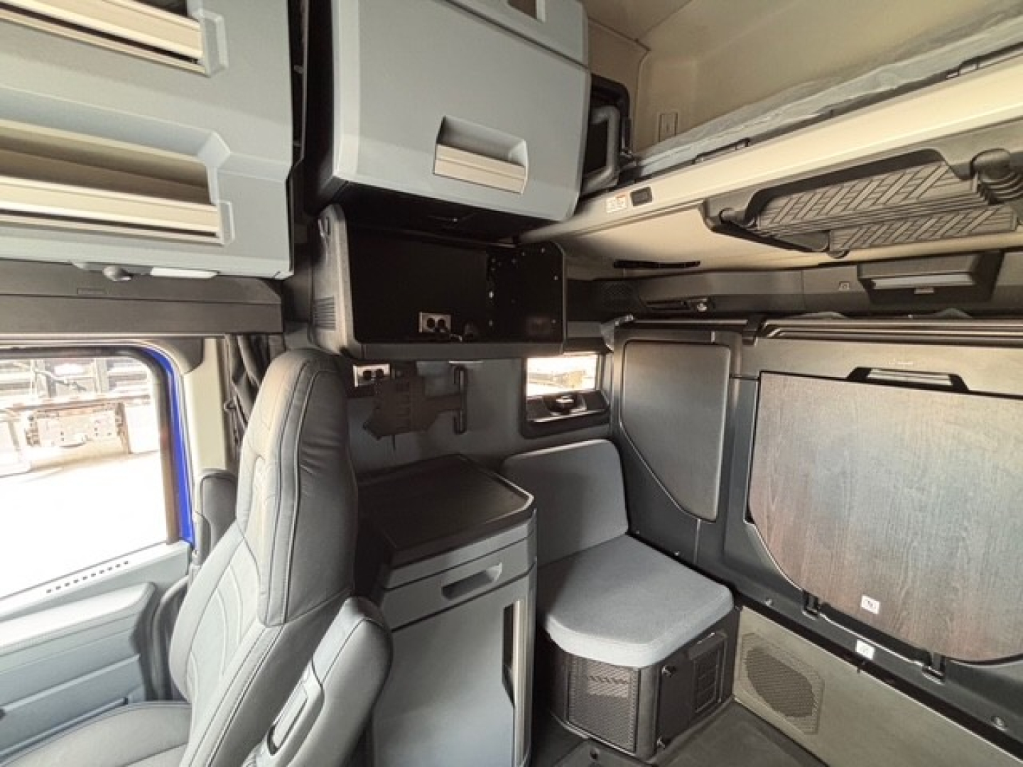 Truck sleeper cab interior showing overhead storage cabinets, a small bench chair, and side paneling with fabric-wrapped walls.