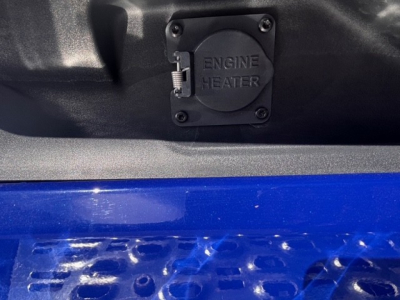 Truck engine bay inlet showing a black engine heater cover with cap and latch in a Mack PR64T76H cab and blue bed panel.