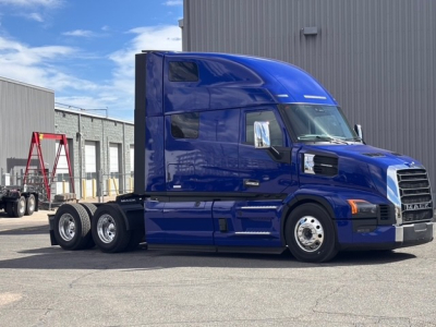 Blue Mack PR64T76H truck tractor with sleeper cab, side view showing chrome fuel tanks and dual rear axles on a dealership lot.
