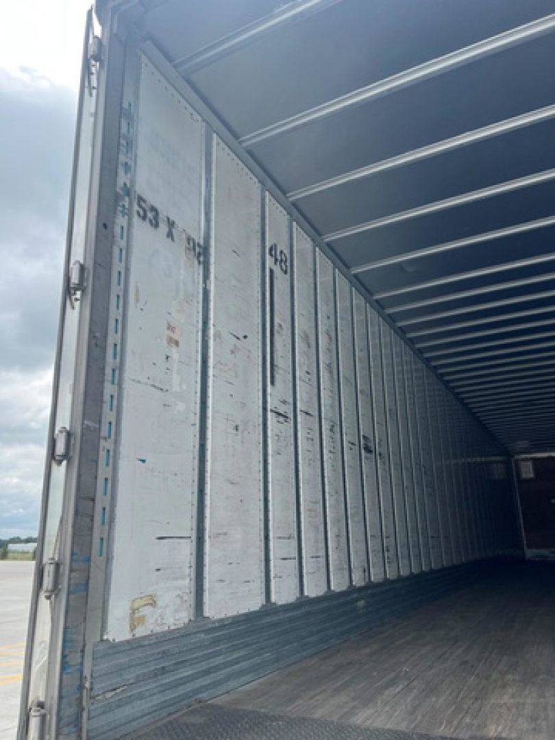 Trailer interior showing metal walls with vertical ribs and wooden floor; open rear doors visible with stamped numbers and markings.